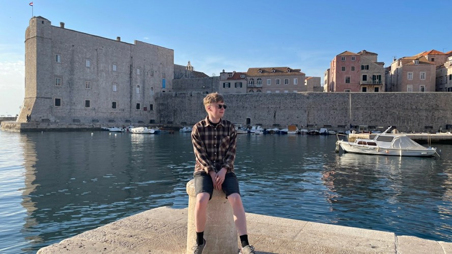 Jere Matias Huttunen istuu ulkomailla sijaitsevassa rantasatamassa. Taustalla on tyyni meri, kivinen linnake ja veneitä.