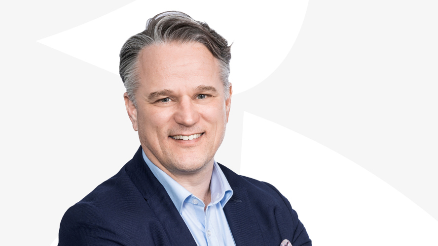 Toimitusjohtaja Janne Vainio katsoo hymyillen kameraan vaaleaa taustaa vasten.