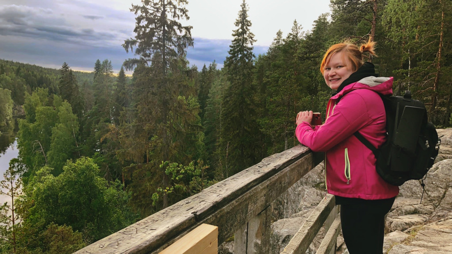 Sovellusasiantuntija Niina Pekkarinen seisoo metsässä näköalatasanteella. Maisemassa pilkottaa järvi metsän keskellä.